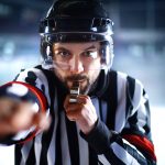 A photo where a hockey referee blows a whistle and points at the viewer.