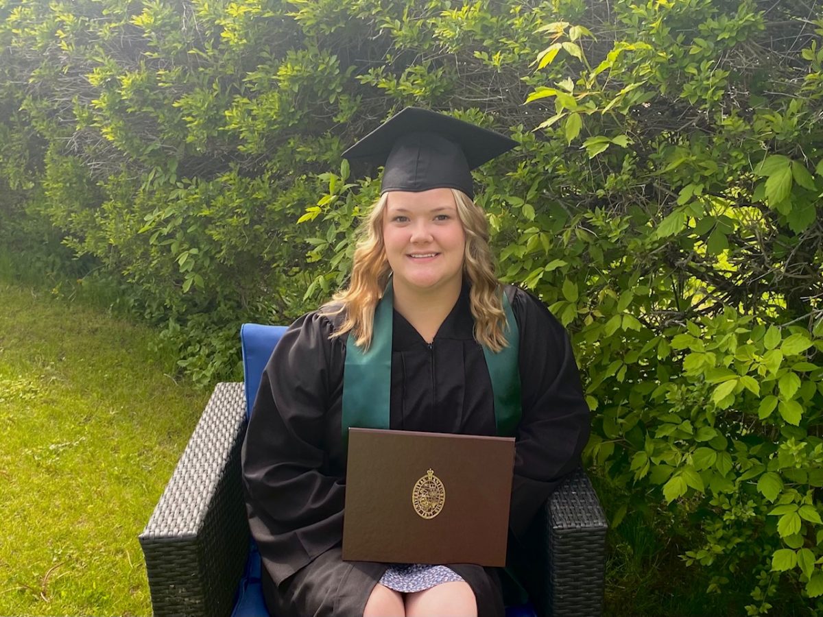 Woman sitting in patio chair, outdoors in front of green trees. She is wearing a grad cap and gown and holding her degree.