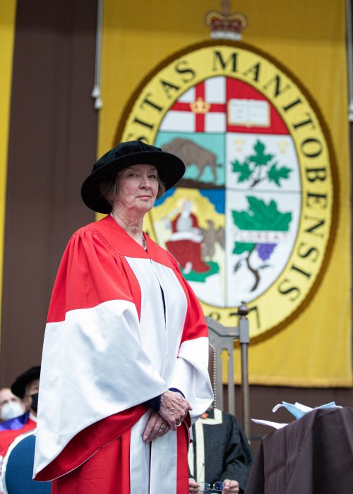 Jan Currie stands in her robes in front of a University of Manitoba banner at convocation.
