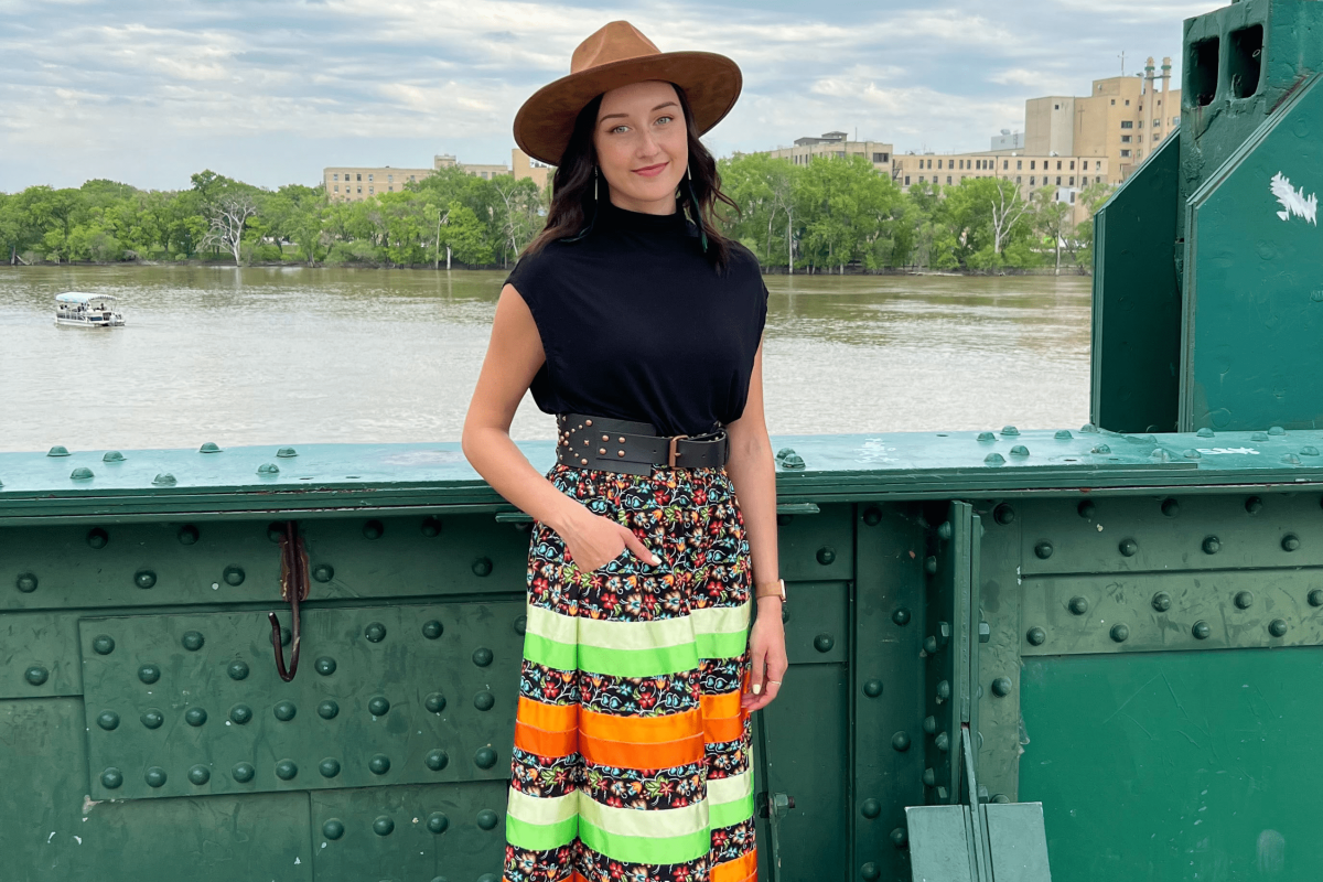 Katie McKenzie wearing a ribbon skirt standing on a green bridge overlooking the Red River.