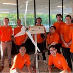 The WE Design team of ten engineering students, each wearing orange shirts and standing alongside their wind turbine.