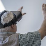 An elderly gentleman wearing a VR headset while sitting in a wheelchair in a nursing home.