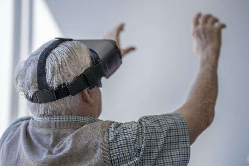 An elderly gentleman wearing a VR headset while sitting in a wheelchair in a nursing home.