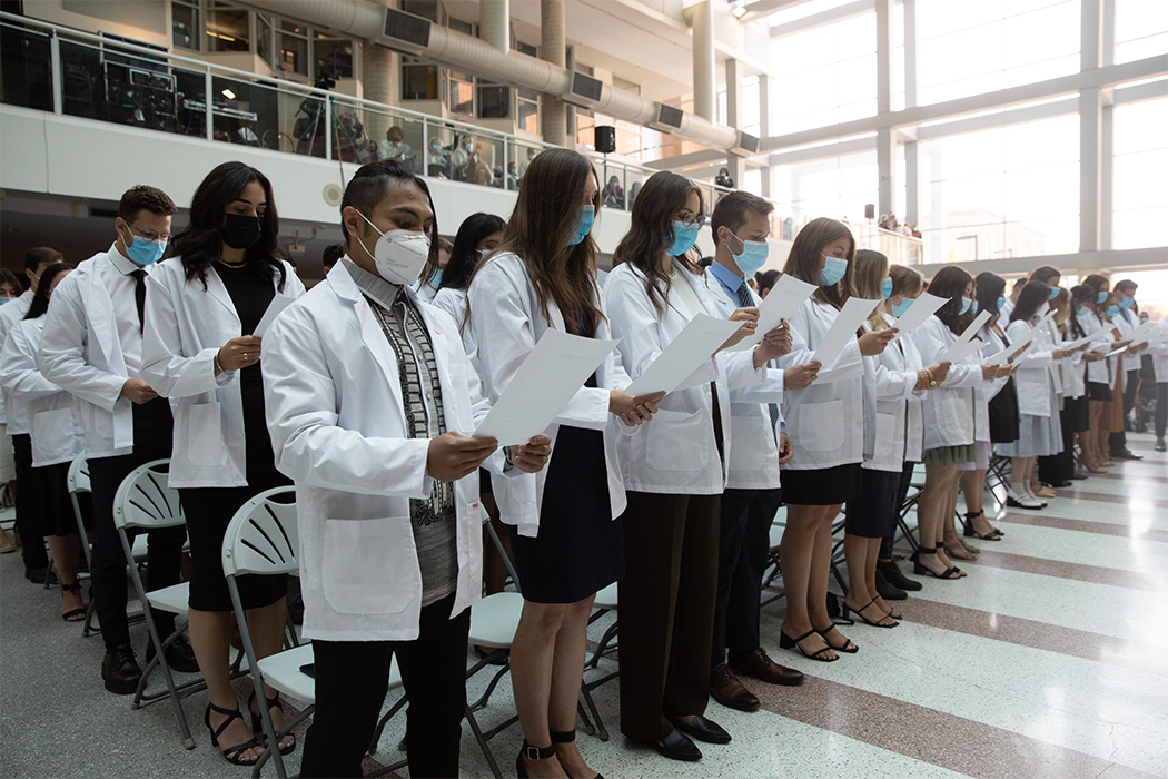 The members of the Medicine Class of 2026, all wearing white coats, stand to recite the Physician's Pledge.