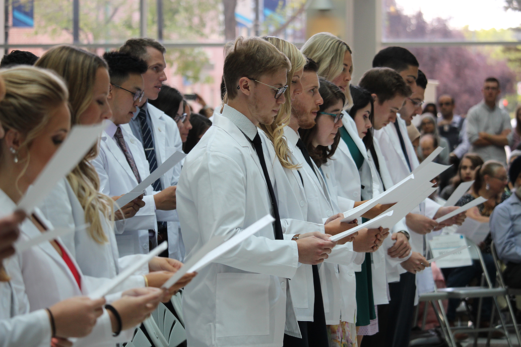 Students wearing white coats each hold a piece of paper. They are reading the Physician's Pledge.