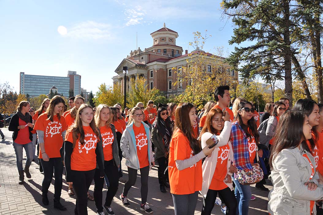A crowd of students, faculty and university staff walk past the University of Manitoba administration building wearing orange shirts.