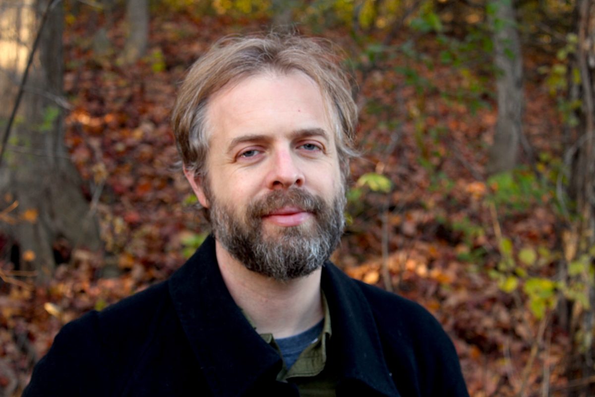 Closeup of man with grey hair and full beard in front of a background of fall leaves.