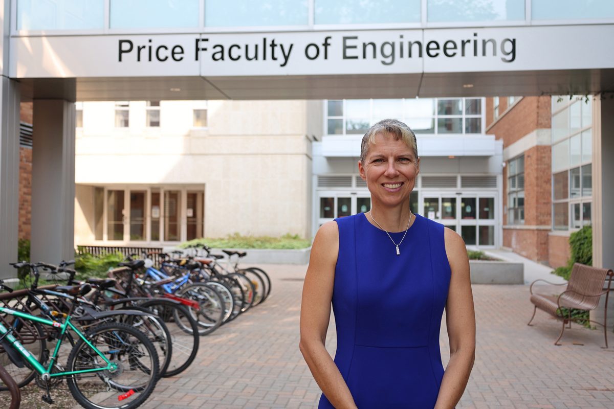 Dr. Marcia Friesen standing, smiling and posting for the camera in front of the E I T C - S P C sky walk. On the silver skywalk in black letters it reads "Price Faculty of Engineering. Marcia is wearing a blue dress. She is outside on a sunny day with a full bike rack in the background.