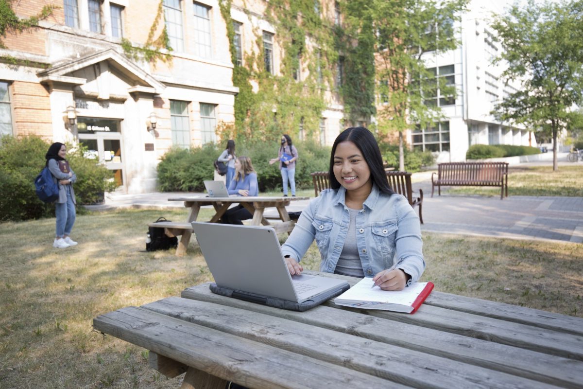 A student works on a laptop outside on a picnic bench. She is smiling as she writes in a notebook