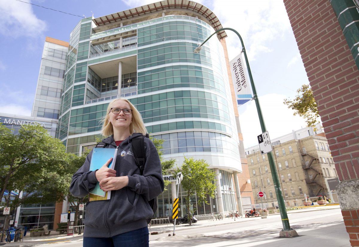 A blonde student with glasses wearing a grey UM hoodie smiles as they look into the distance. They are standing in front of Brodie Centre on UM's Bannatyne Campus.