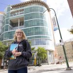 A blonde student with glasses wearing a grey UM hoodie smiles as they look into the distance. They are standing in front of Brodie Centre on UM's Bannatyne Campus.