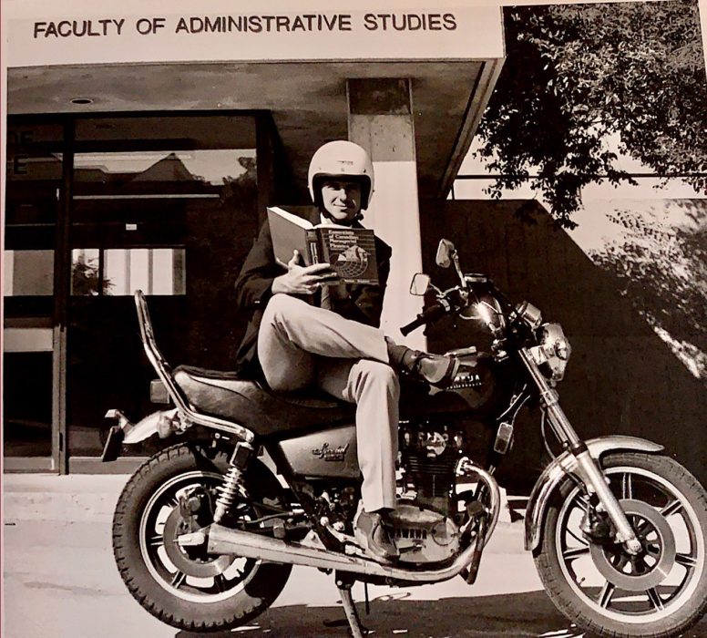An old black and white photo of Professor John McCallum, sitting on a motorcycle reading a book outside the University of Manitoba Faculty of Administrative Studies building.