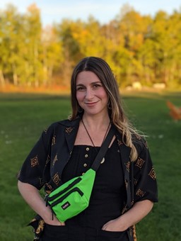 Eliana Hotz wears a black outfit with a neon green bag. They are standing on the edge of a grassy field. The late day sun shines on trees in the background.