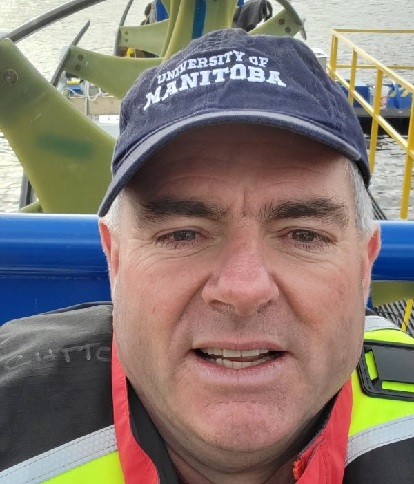 Eric Bibeau wearing a life jacket on a small boat alongside the rivgen turbine. He is smiling for the camera. 