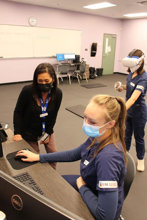 A student and instructor look at a computer screen while another student works behind them wearing a virtual reality headset and hand controls. 