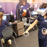 A nursing student wears a virtual reality headset and hand controls while another student works at a computer station behind her.