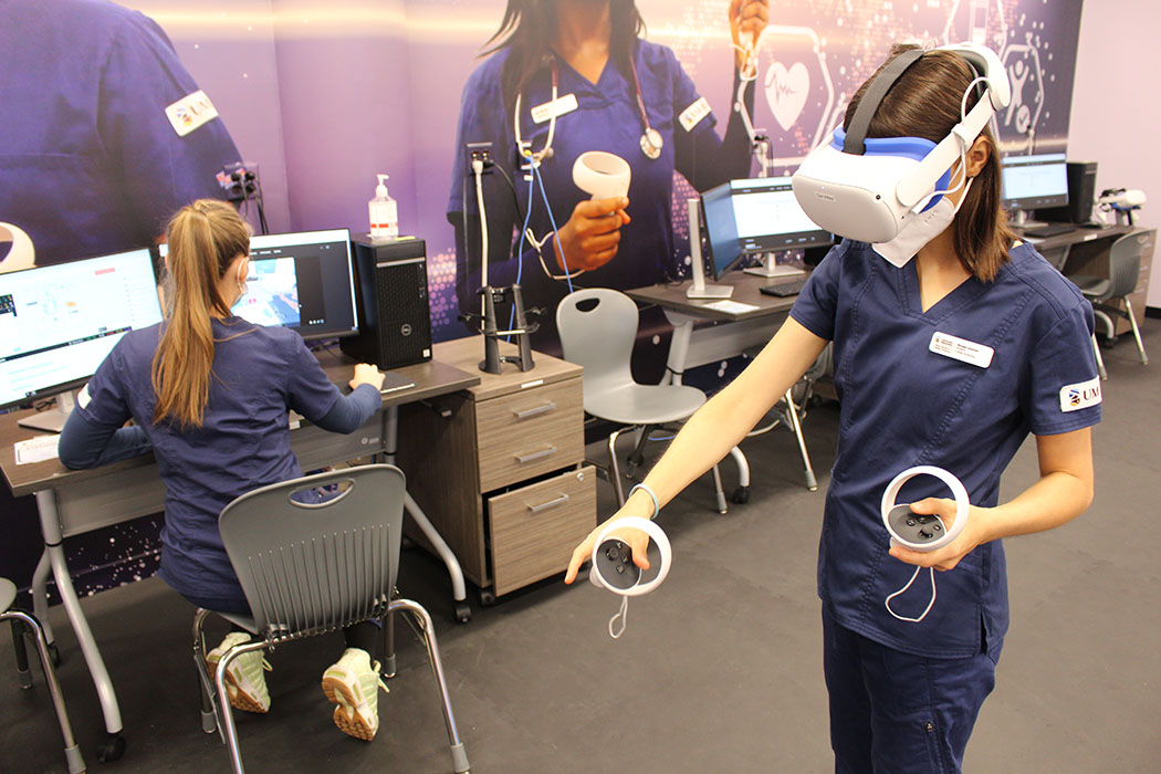 A nursing student wears a virtual reality headset and hand controls while another student works at a computer station behind her.