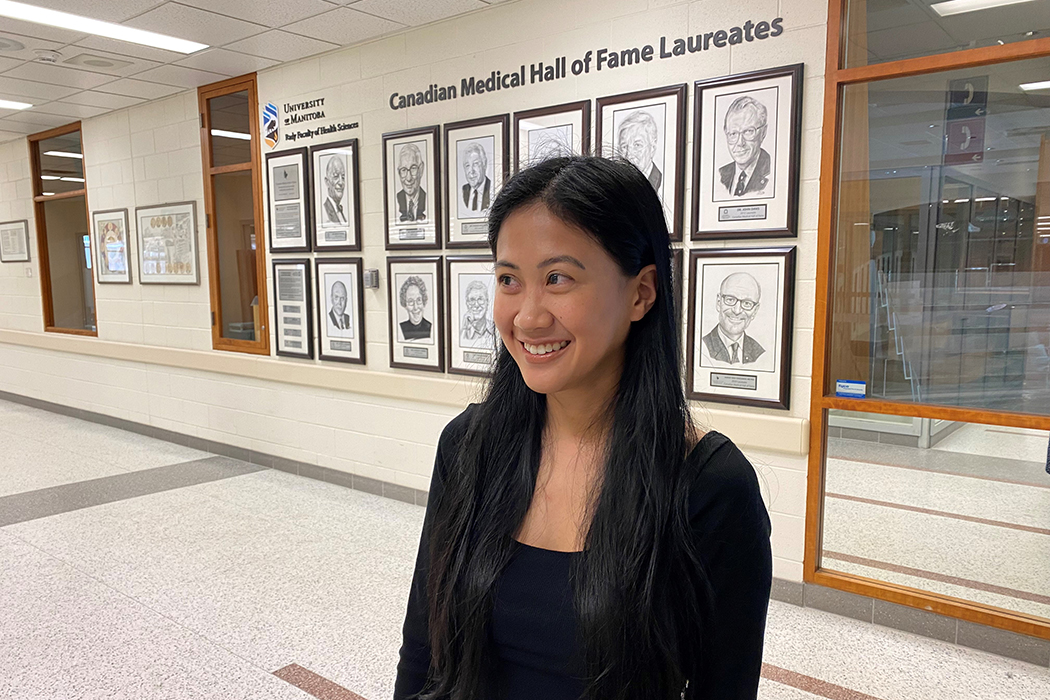 Valcourt poses for photo in front of 12 framed drawings of the CMHF laureates. Text on the wall reads "Canadian Medical Hall of Fame Laureates."