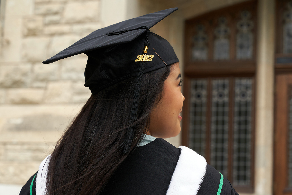 A UM grad in cap and down looks off into the distance. Their tassle has 2022 on it and they stand in front of a limestone brick building with stained glass doors.