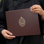 A graduating student wearing a black gown holds a UM parchment folder in their hands. It is brown with gold accents
