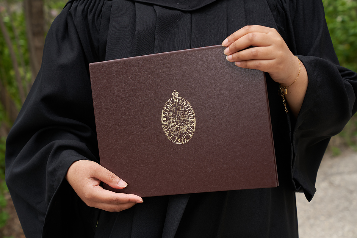 A graduating student wearing a black gown holds a UM parchment folder in their hands. It is brown with gold accents