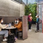 Three students study together in a booth. Two other students stand chatting in the background. They are all wearing KN-95 masks.