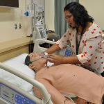 A nursing instructor works with a life-sized manikin in a simulated hospital setting.