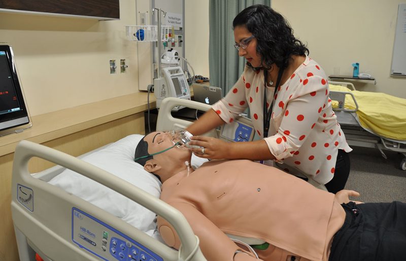 A nursing instructor works with a life-sized manikin in a simulated hospital setting.