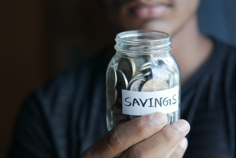 A person holds a glass jar full of coins marked as "savings"