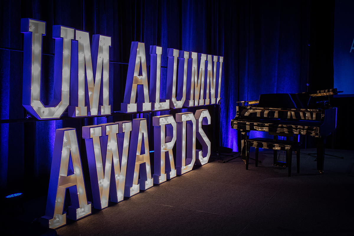 View of a stage with large letters that speel out UM Alumni Awards with a grand piano in the background.