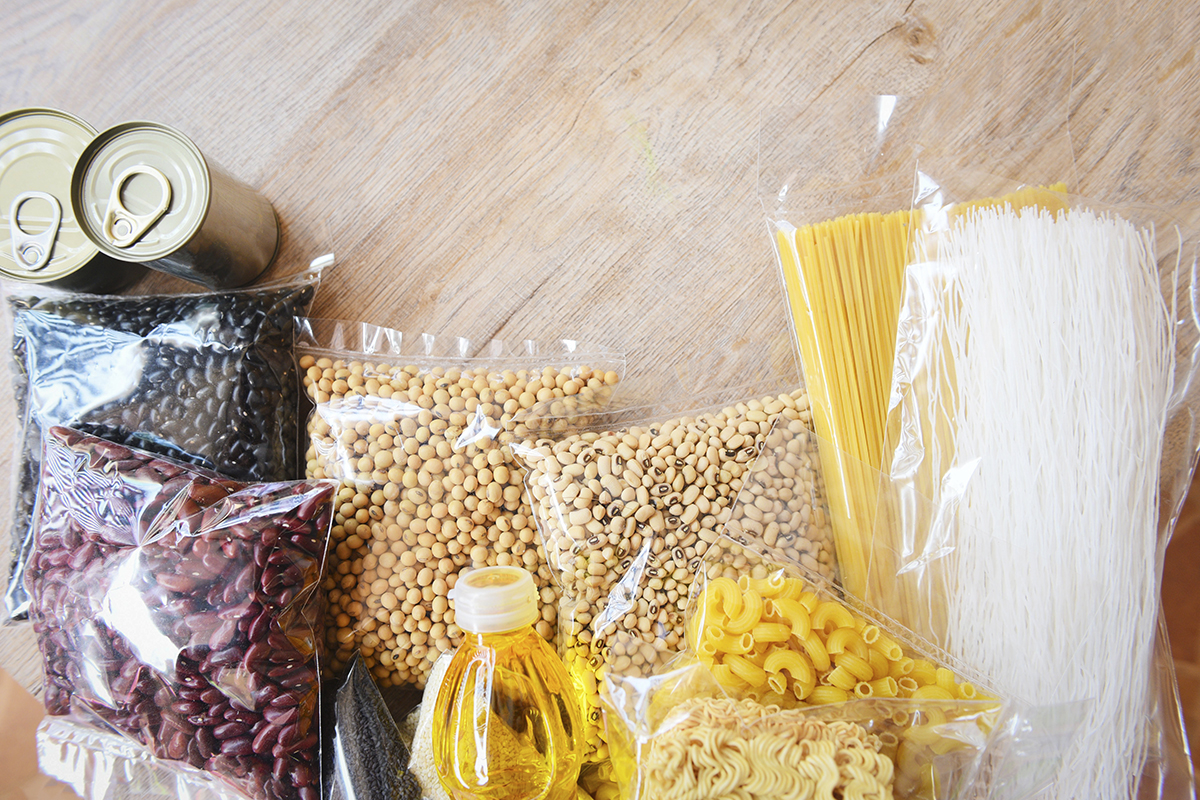 Unbranded canned food, bags of beans and pasta and bottle of cooking oil lay spread out on a wooden table