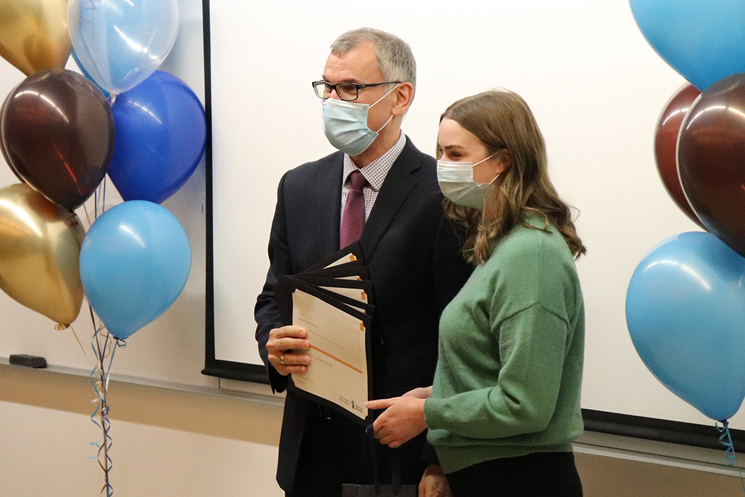 Dr. Vercaigne and Shaelyn pose for a photo. Dr. Vercaigne is holding five certificates. Balloons in UM's colours are on either side of them.