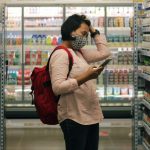 A person wearing a mask and a red backpack stands in the aisle of a grocery store. They hold their phone in one hand as their other hand runs through their hair. They look stressed.