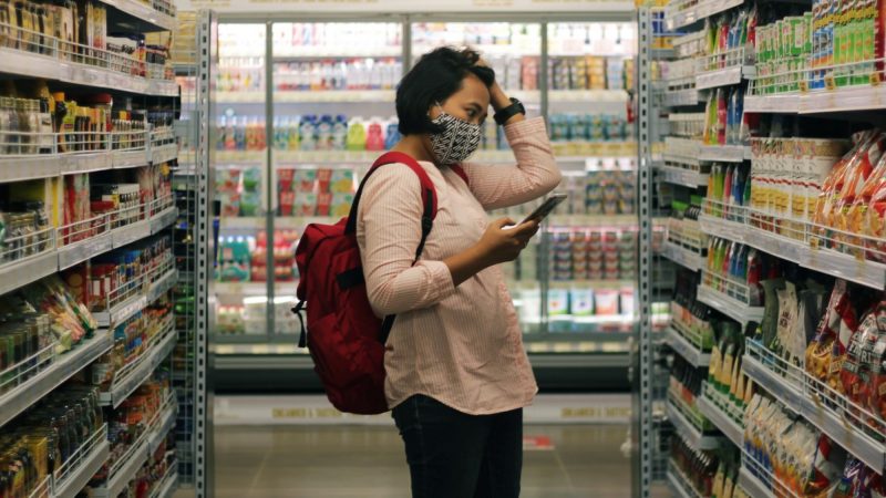A person wearing a mask and a red backpack stands in the aisle of a grocery store. They hold their phone in one hand as their other hand runs through their hair. They look stressed.