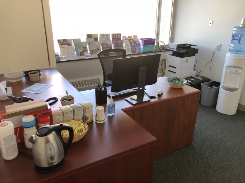 The view when someone arrives at the Sexual Violence Resource Centre. A desk with a computer, a kettle and a variety of teas. A row of pamphlets lines the window sill behind the desk. 