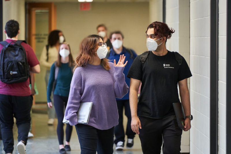 Two students featured walking together indoors, while masked., various other students, also masked are walking by.