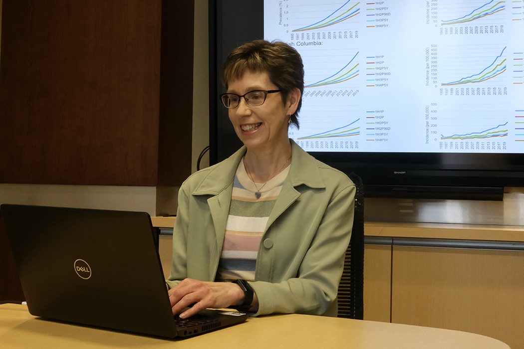 Dr. Lisa Lix sits at a table and uses a laptop. On a screen behind her are six graphs.