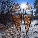 Traditional snowshoes sit in the foreground of a blue sky on a winter day.