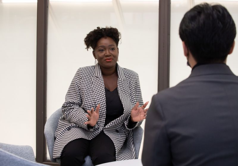 Carine sits in a suit jacket speaking to a student. The student is facing Carine.