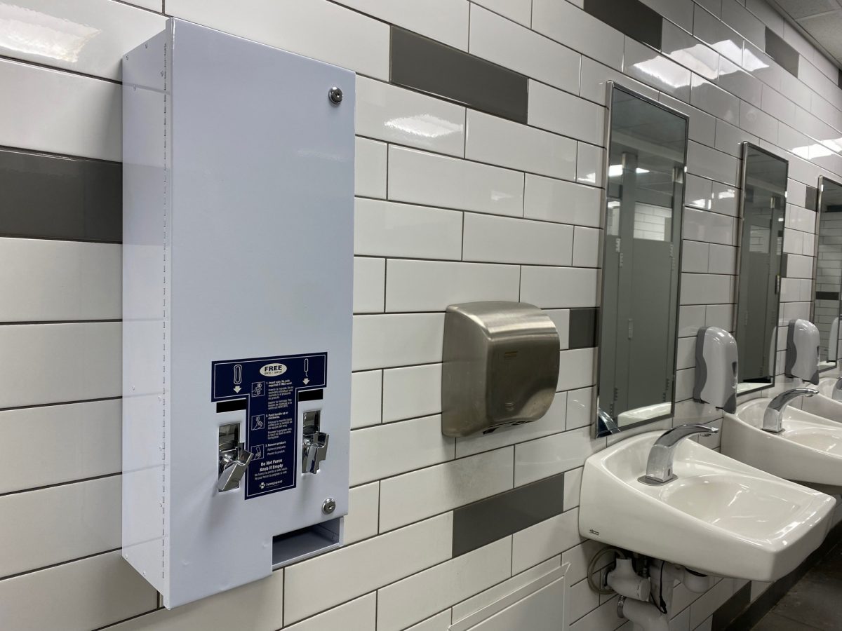 Menstual product dispenser affixed to white-tiled bathroom wall next to air hand driver and a row of sinks.
