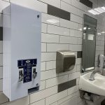 Menstual product dispenser affixed to white-tiled bathroom wall next to air hand driver and a row of sinks.