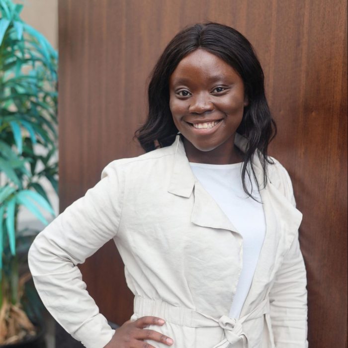 Clèche Kokolo smiles in front of a brown office door. She's wearing white and a green plant catches light in the background.