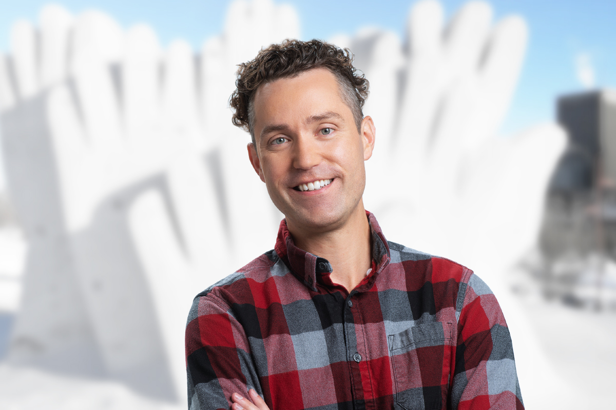 Man in plaid shirt with arms crossed, a snow sculpture behind him.