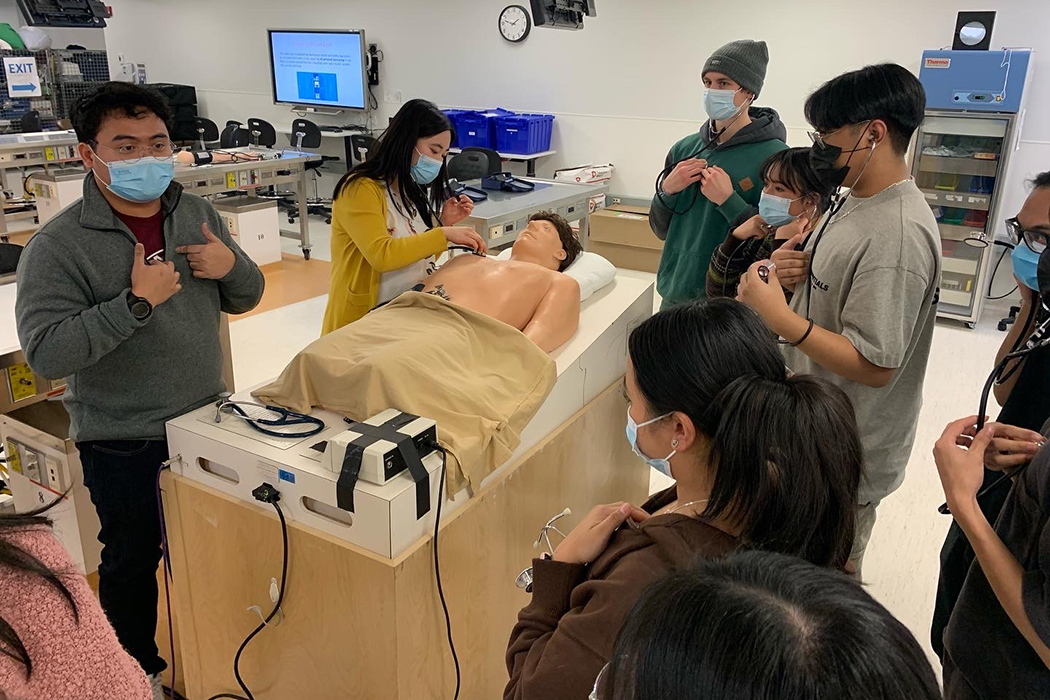 Students wearing stethoscopes gather around instructor. One person places stethoscope on chest of training manikin.