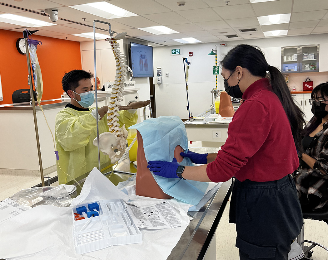 A student places her hands on a training model of a back. The instructor has his hands extended as he explains a step in the procedure. A model of a spine is on the table.