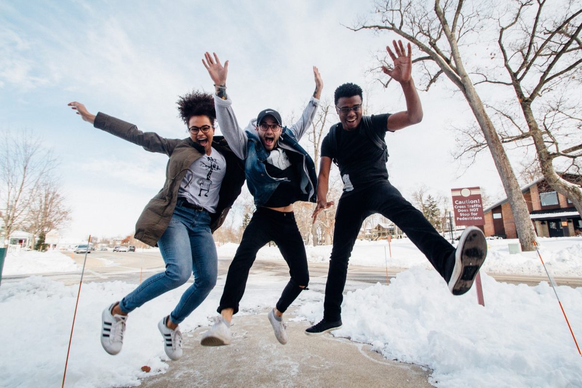 Three happy students jump in the air and are captured by the camera mid-jump. They are on a concrete sidewalk cleared of snow and bare trees are in the background.