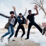Three happy students jump in the air and are captured by the camera mid-jump. They are on a concrete sidewalk cleared of snow and bare trees are in the background.