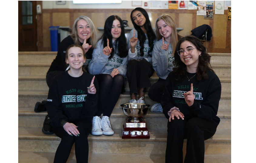 Five students and 1 teacher sitting around a trophy all showing the number 1 winning finger.