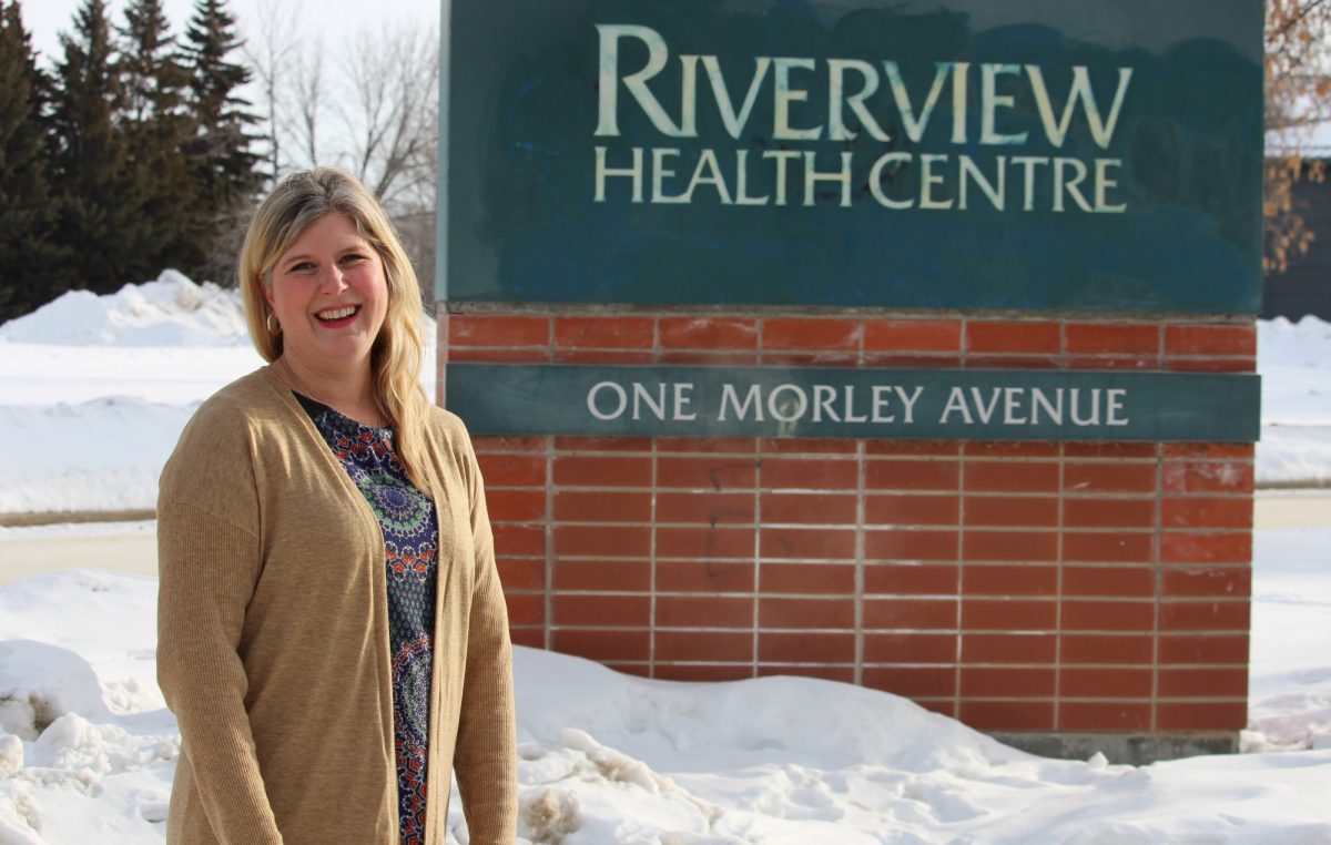 Genevieve Thompson stands outside by a sign that reads "Riverview Health Centre."