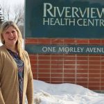 Genevieve Thompson stands outside by a sign that reads "Riverview Health Centre."